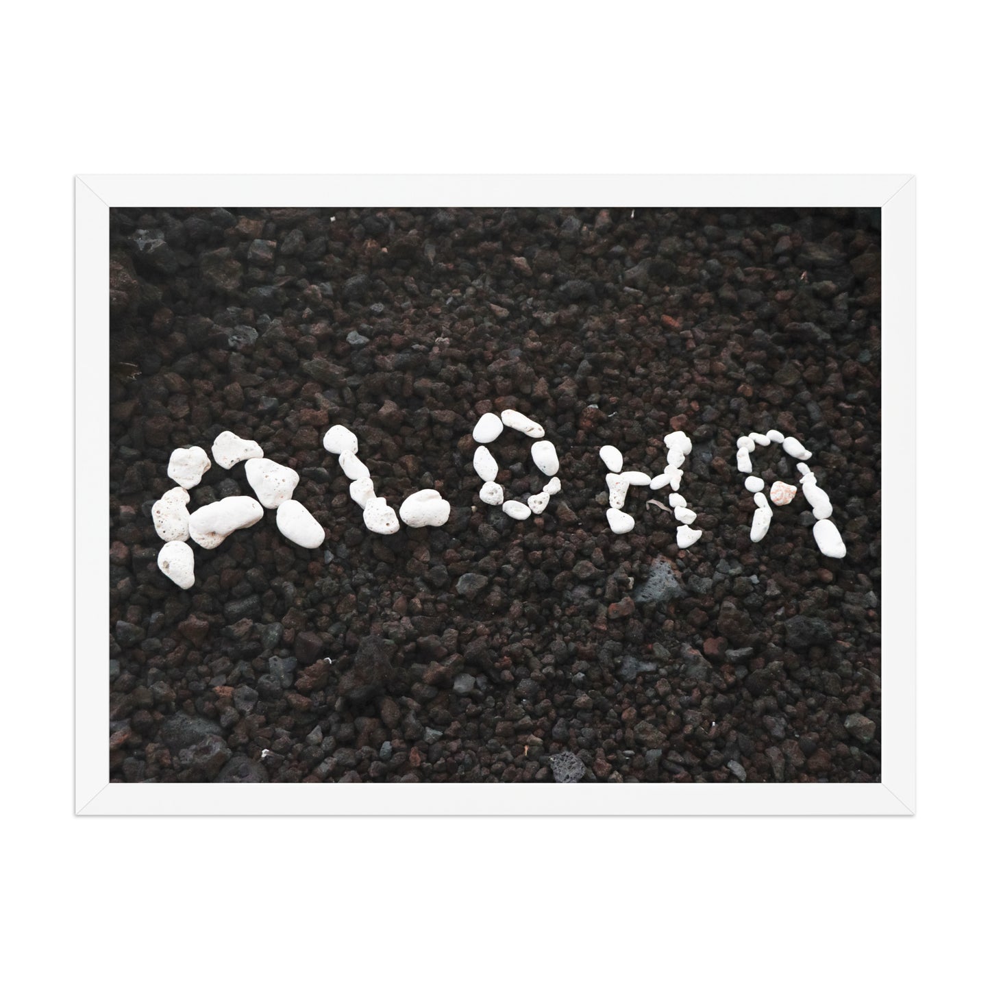 White stones spell Aloha against dark beach sand in a simple, graphic coastal composition; white frame, 18 × 24 inches.