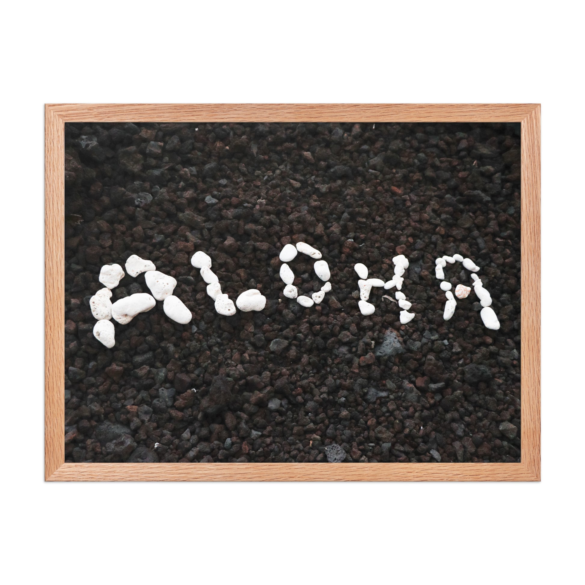 White stones spell Aloha against dark beach sand in a simple, graphic coastal composition; wood frame, 18 × 24 inches.