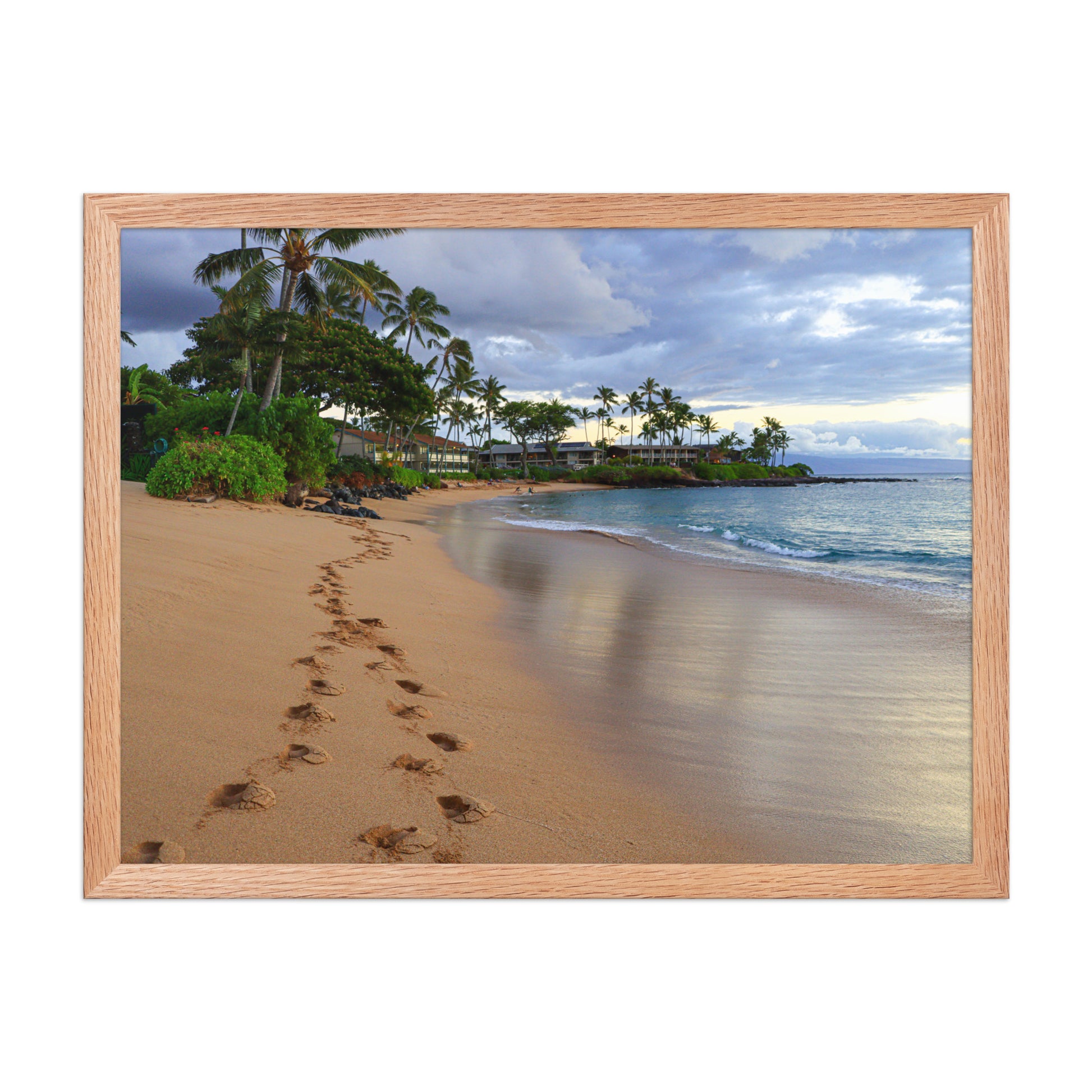 A trail of footprints crosses a quiet sandy beach beside gentle water and coastal vegetation; wood frame, 18 × 24 inches.