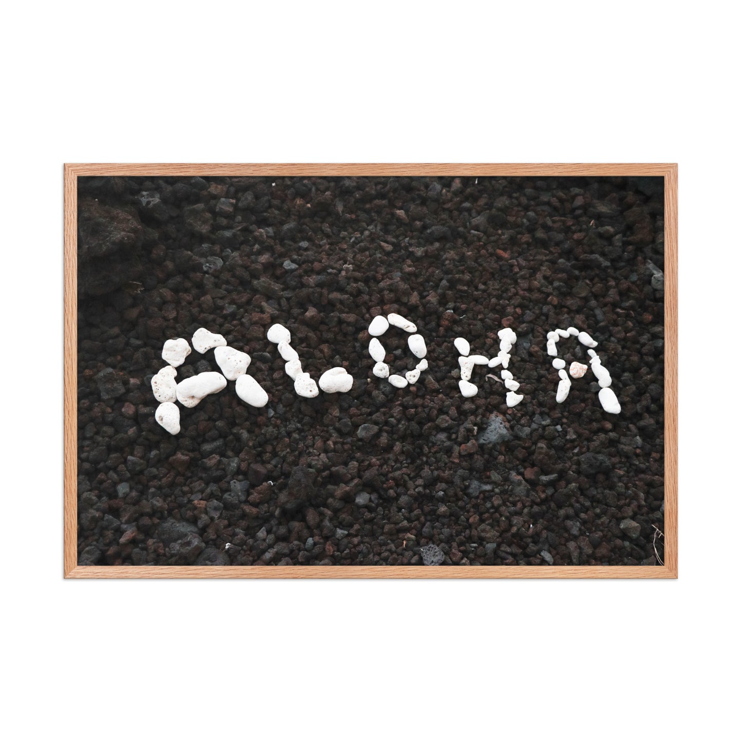 White stones spell Aloha against dark beach sand in a simple, graphic coastal composition; wood frame, 24 × 36 inches.