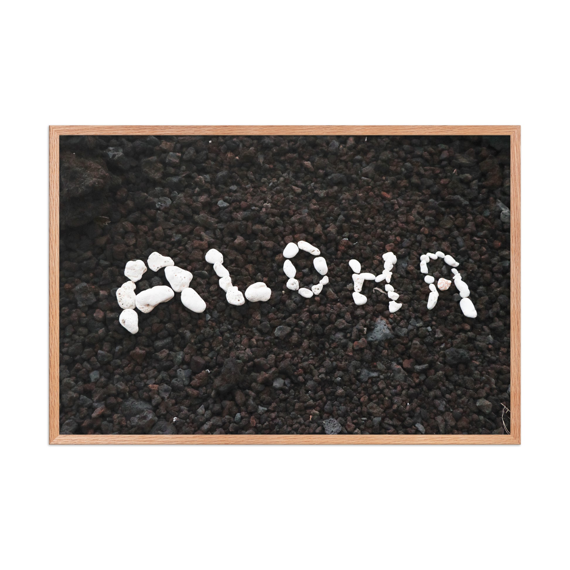 White stones spell Aloha against dark beach sand in a simple, graphic coastal composition; wood frame, 24 × 36 inches.
