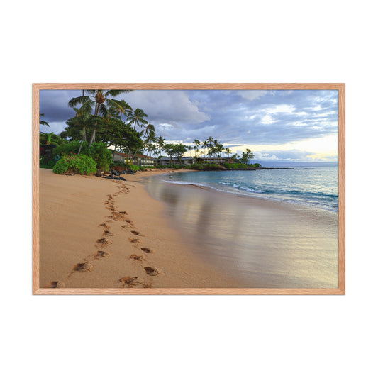 A trail of footprints crosses a quiet sandy beach beside gentle water and coastal vegetation; wood frame, 24 × 36 inches.