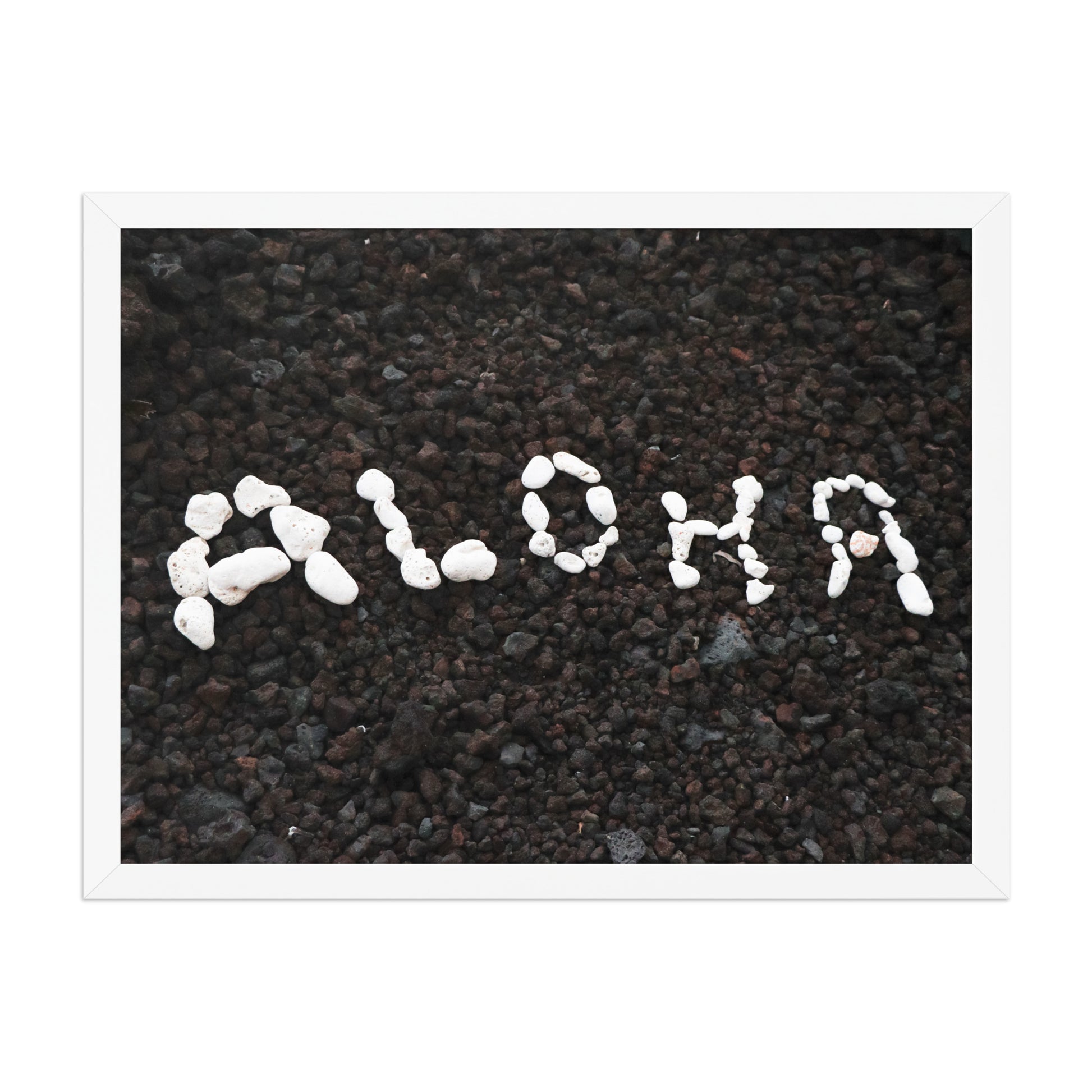 White stones spell Aloha against dark beach sand in a simple, graphic coastal composition; white frame, 18 × 24 inches.