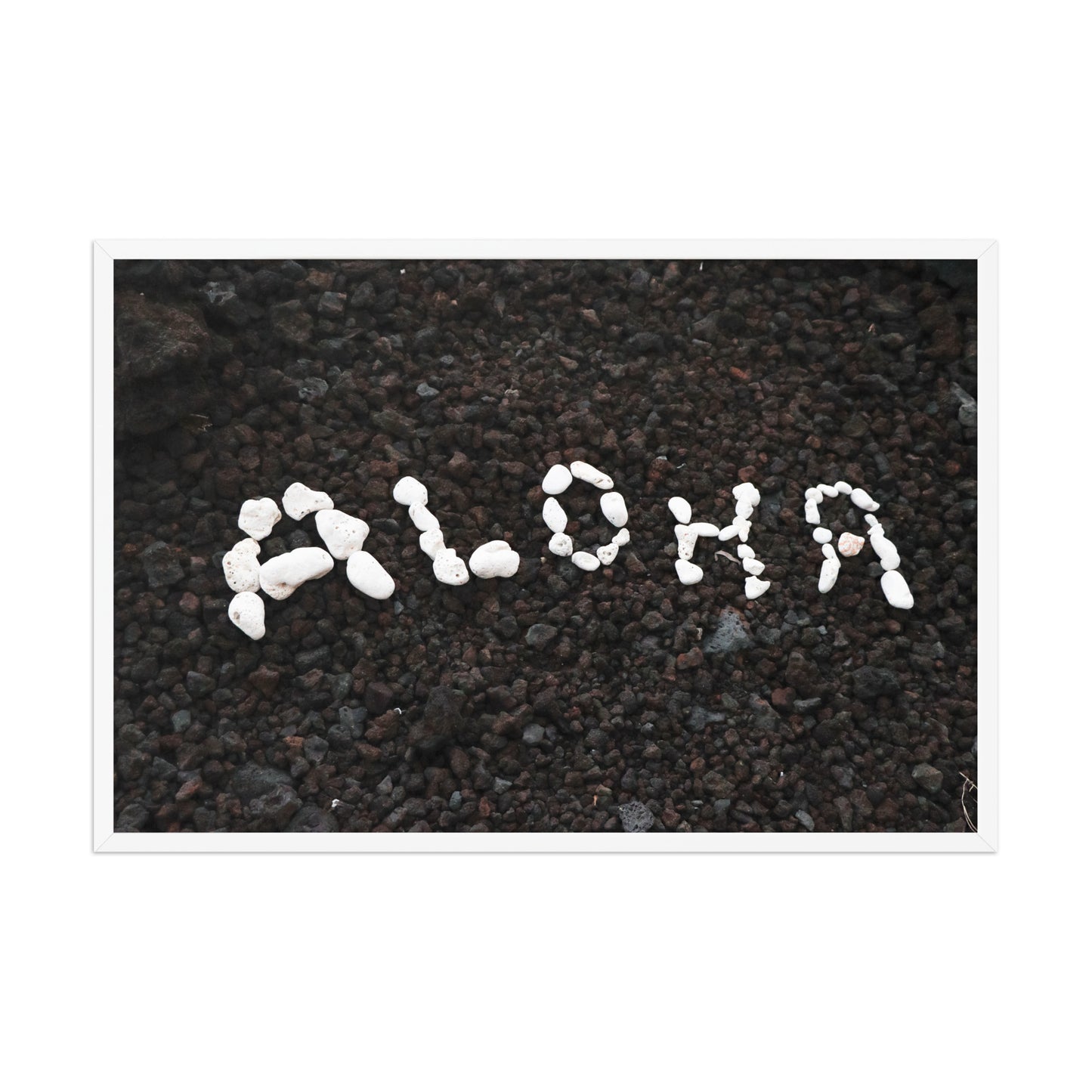 White stones spell Aloha against dark beach sand in a simple, graphic coastal composition; white frame, 24 × 36 inches.