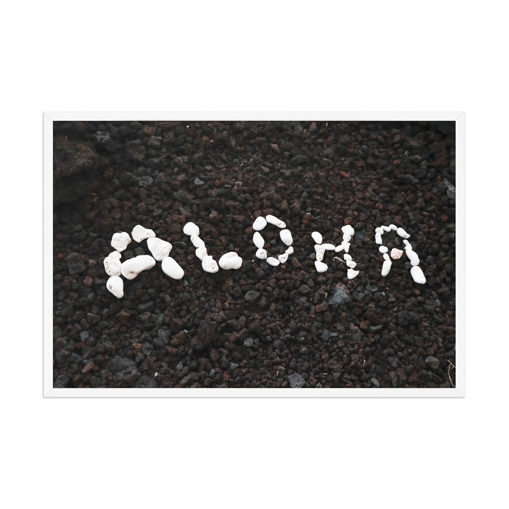 White stones spell Aloha against dark beach sand in a simple, graphic coastal composition; white frame, 24 × 36 inches.