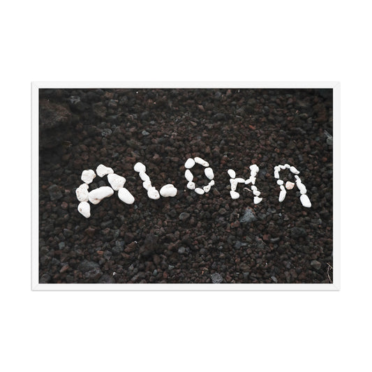 White stones spell Aloha against dark beach sand in a simple, graphic coastal composition; white frame, 24 × 36 inches.
