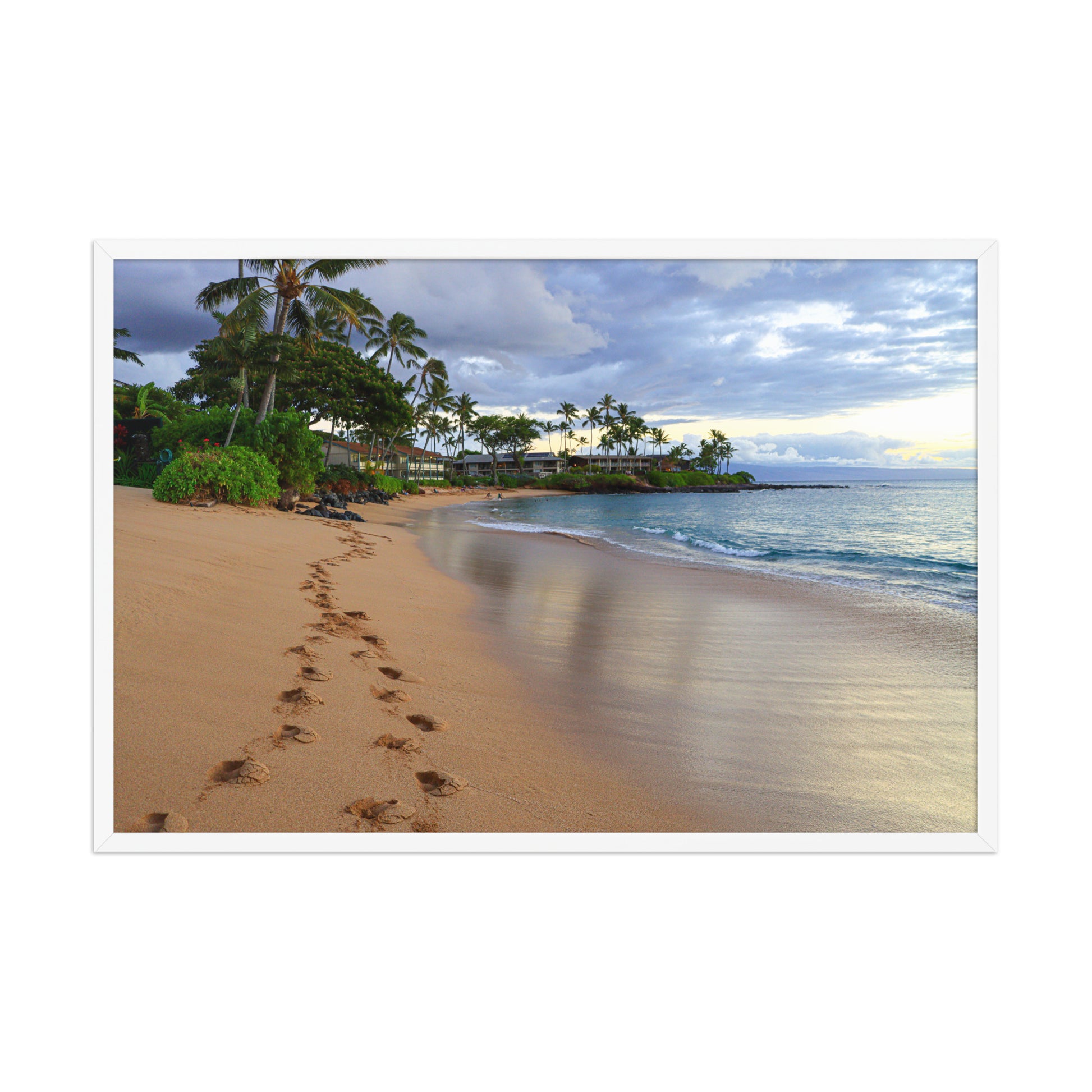 A trail of footprints crosses a quiet sandy beach beside gentle water and coastal vegetation; white frame, 24 × 36 inches.