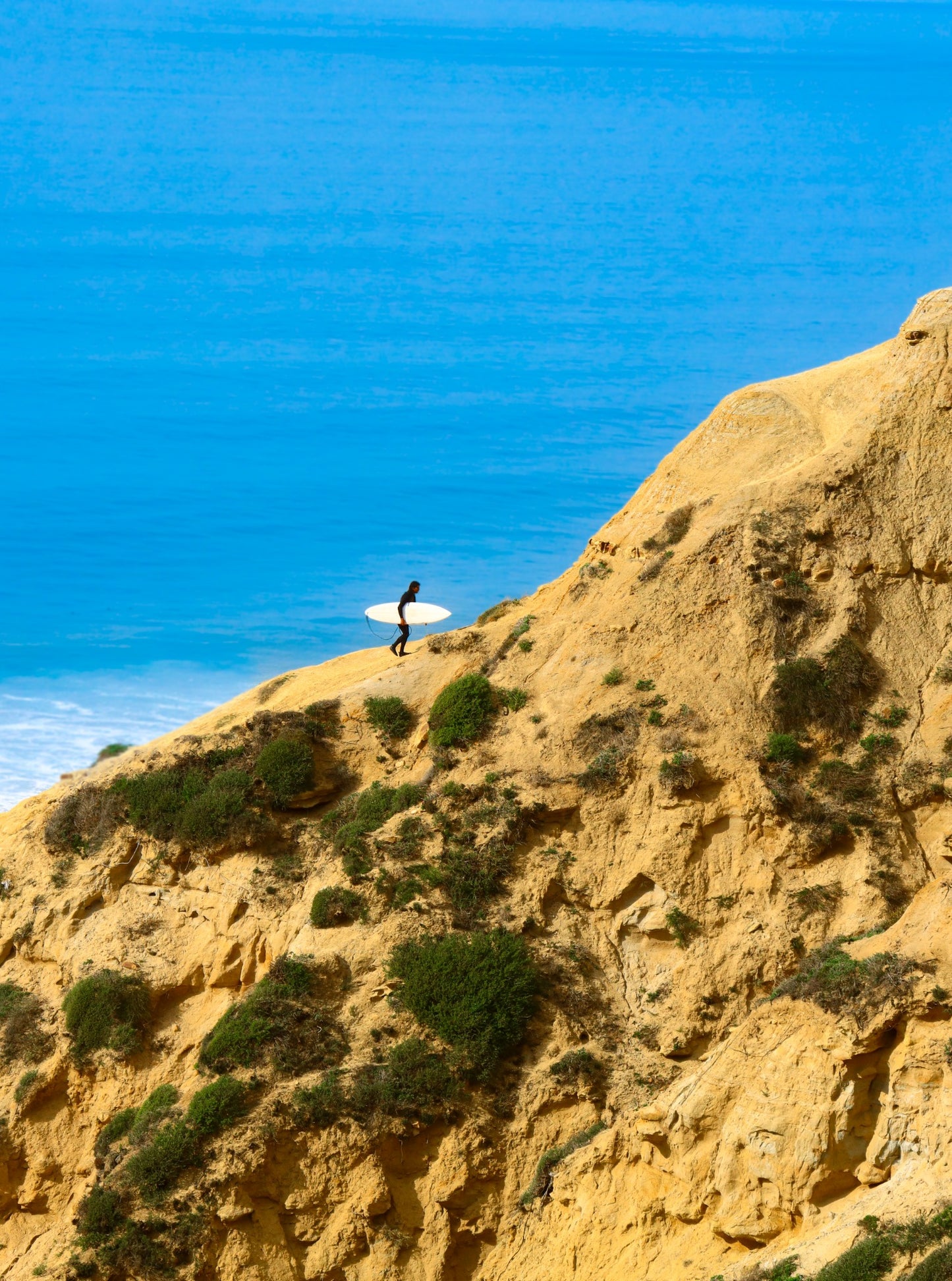 Surfer carrying a white surfboard along a sandstone cliff above blue ocean water.