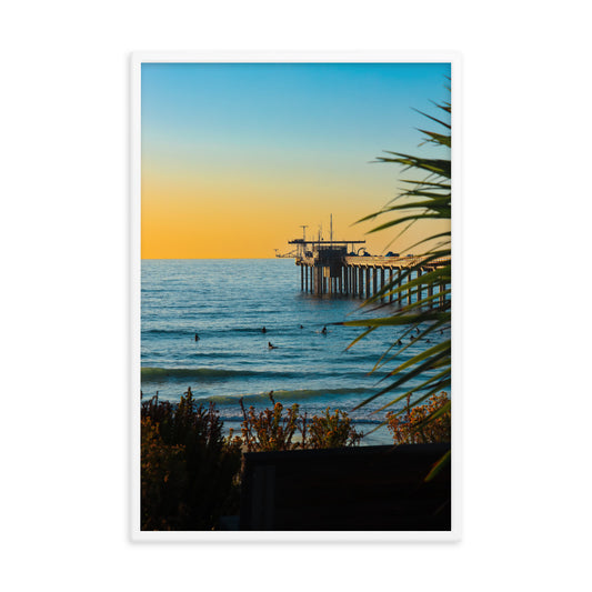 Scripps Pier reaches into the Pacific beneath a soft sunset sky, framed by coastal foliage in La Jolla; white frame, 24 × 36 inches.